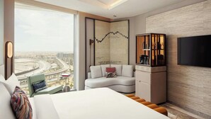 Quarto de hotel moderno com uma cama grande, área de estar com sofá e poltronas, televisão e vista para a cidade.