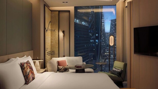 Quarto de hotel moderno com uma cama grande, área de estar, televisão e vista para a paisagem urbana.