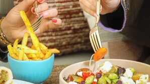 Uma pessoa segurando um garfo com um pedaço de comida, sentada à mesa com uma tigela de batatas fritas e um prato de salada.