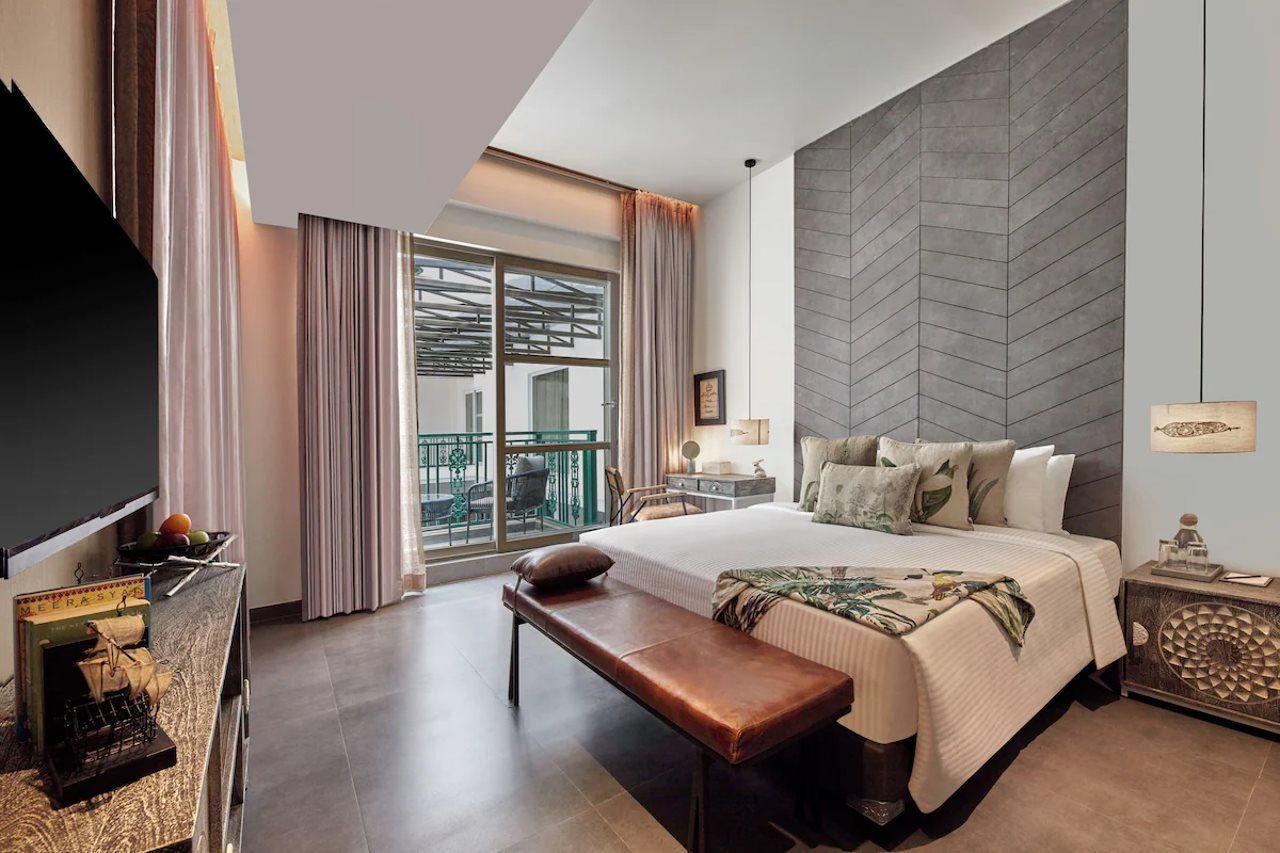Ein Hotelzimmer mit einem großen Bett, einer Bank, einem Schreibtisch mit Lampe, einem Fernseher und Blick auf den Balkon.
