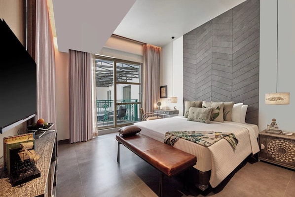 Habitación de hotel con una cama grande, un banco, un escritorio con lámpara, un televisor y vistas al balcón exterior.