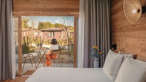 A bedroom with a large bed, wooden walls, a view of a garden, and a dining area with a table and chairs.