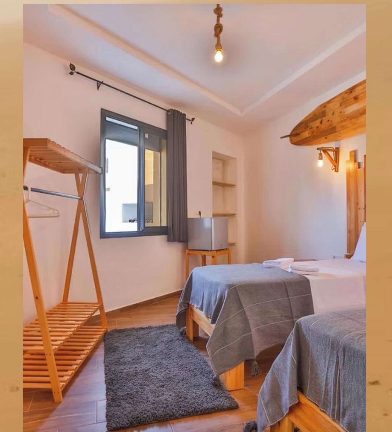 A room with two beds, a wooden shelf, a small table, a mirror, and a window with curtains.