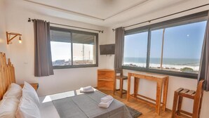 A bedroom with a bed, a desk, a TV, and large windows with a view of the sea. - Moon surf (Imsouane mroc)