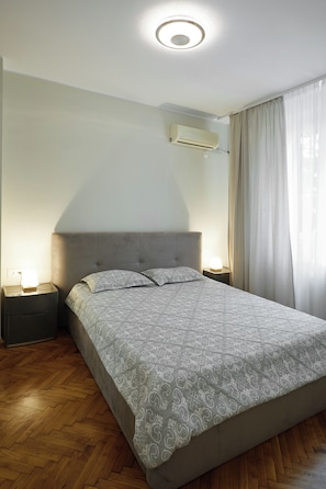 Ein Schlafzimmer mit einem Bett, Nachttischen, einer Klimaanlage und einem Fenster mit Vorhängen.