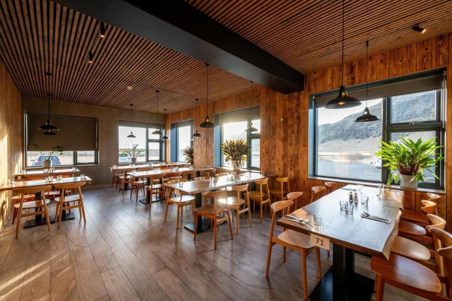 A restaurant interior with wooden floors, tables, and chairs, large windows with a view, and hanging pendant lights.