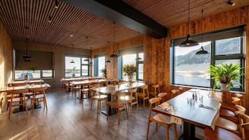 A restaurant interior with wooden floors, tables, and chairs, large windows with a view, and hanging pendant lights.