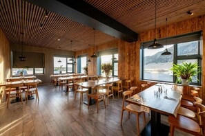 A restaurant with wooden floors, tables, and chairs, large windows with a view, and hanging pendant lights.