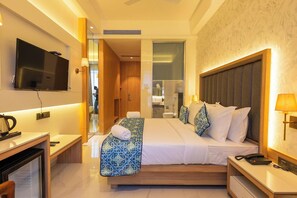 Habitación de hotel con cama, mesita de noche, lámpara, televisión y vista al baño.