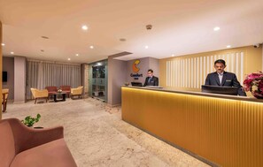A hotel reception area with a reception desk, two receptionists, a seating area with chairs, and a large window with curtains. - Comfort Inn Grand (Ghaziabad)