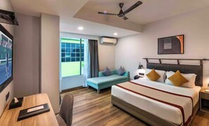 A hotel room with a bed, a desk, a chair, a window with curtains, a ceiling fan, and a mounted picture. - Comfort Inn Grand (Ghaziabad)