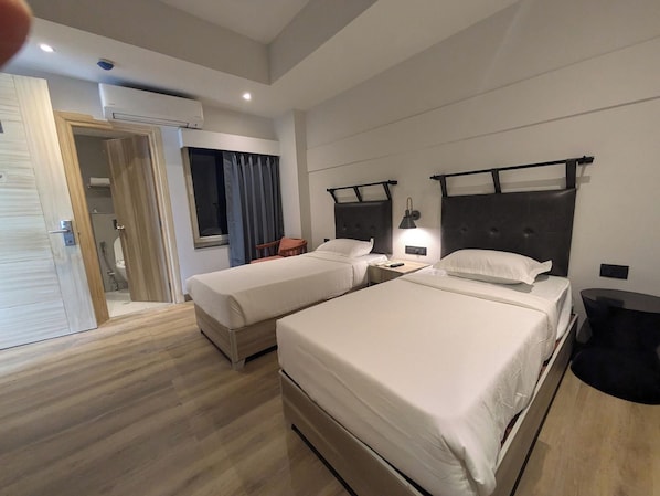 A hotel room with two beds, a wooden floor, a bathroom with a shower, and a window with curtains. - Comfort Inn Grand (Ghaziabad)