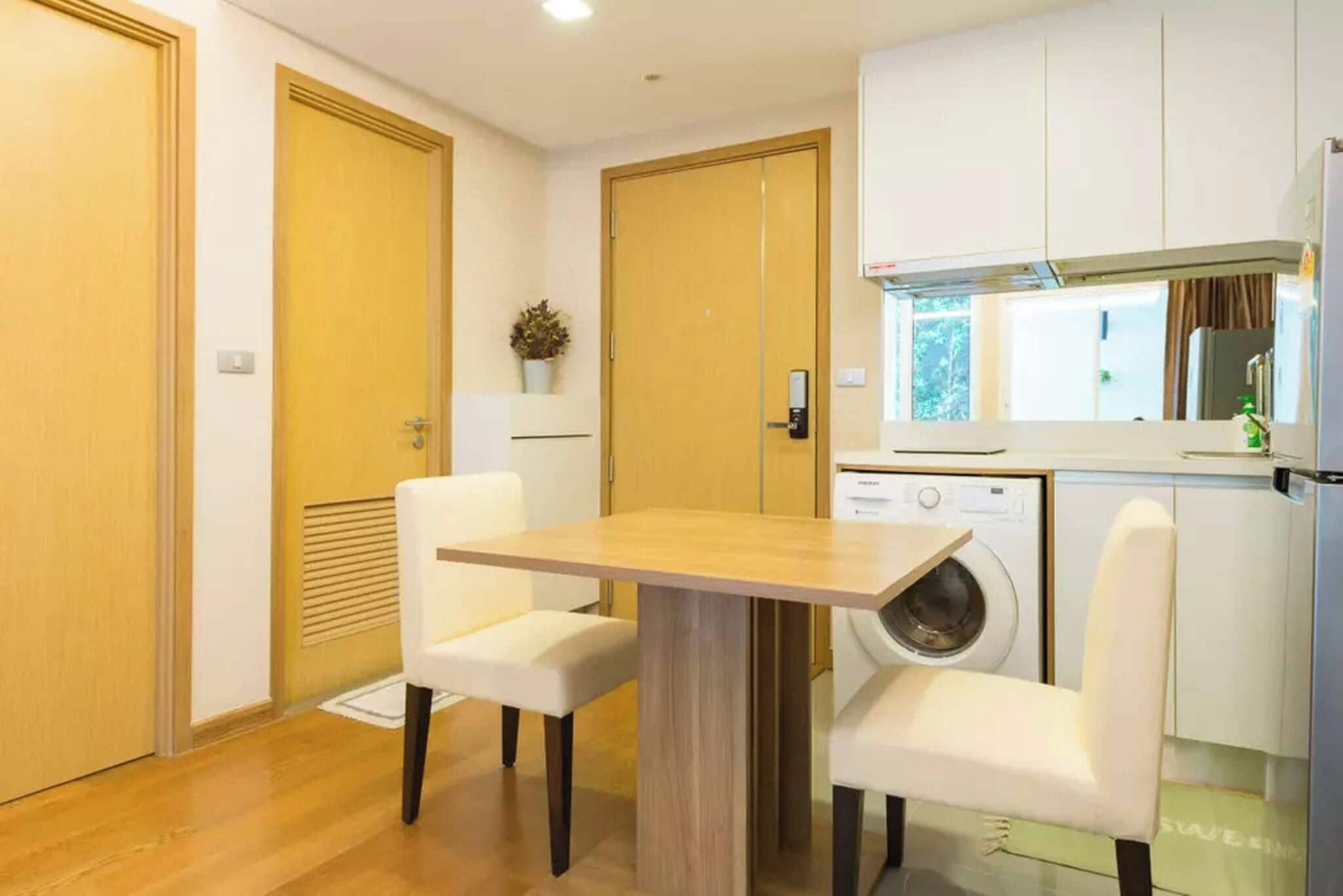 A compact kitchen with a washing machine, a dining table with chairs, and wooden doors.