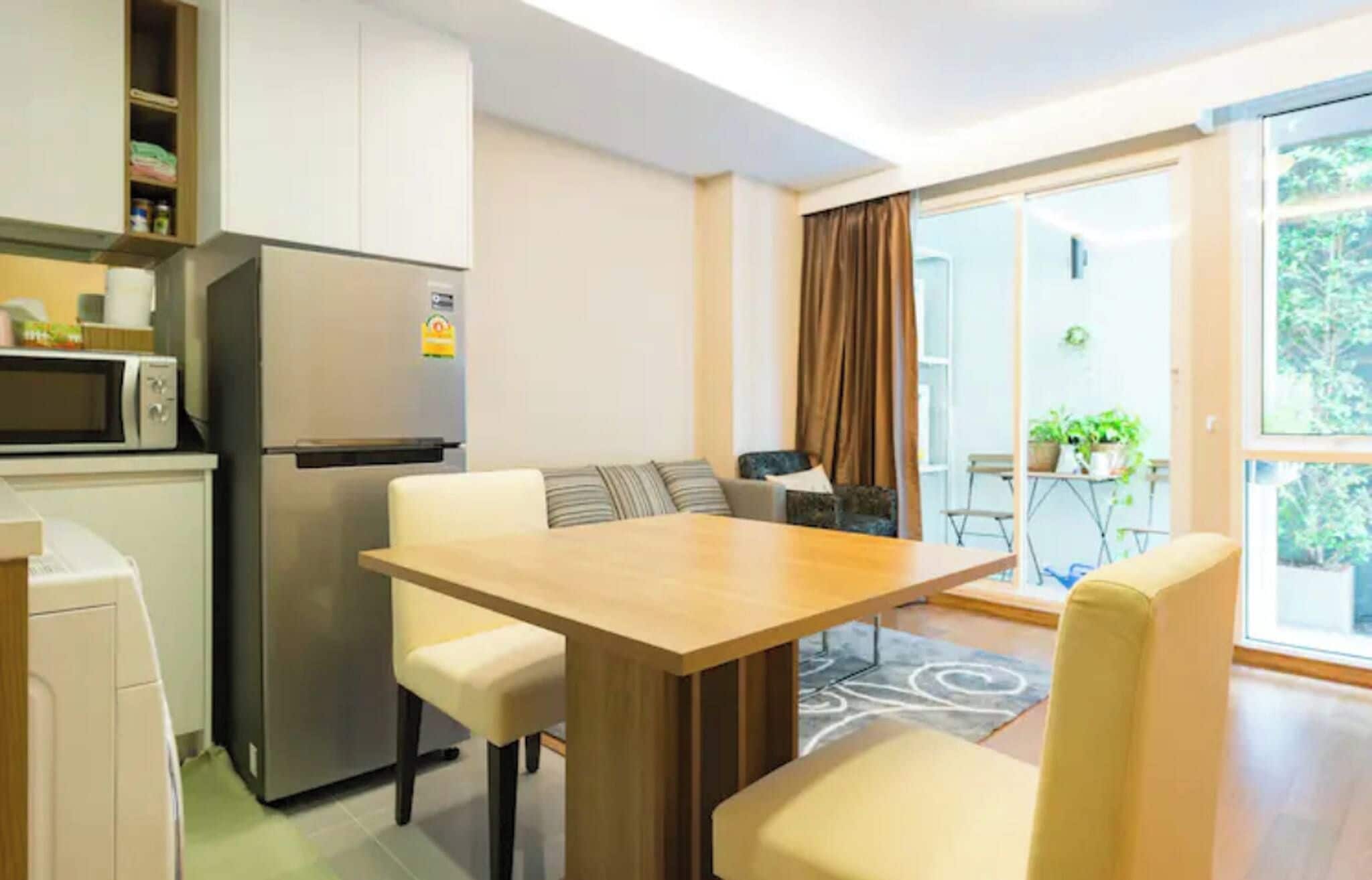 A compact kitchen with a refrigerator, microwave, and cabinets. There is a dining table with chairs and a sofa in the living area. A sliding glass door leads to an outdoor area with potted plants.