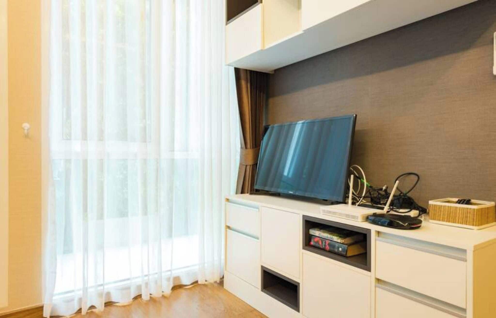 A modern living room with a flat-screen TV on a white entertainment unit, a large window with sheer curtains, and a wooden floor.
