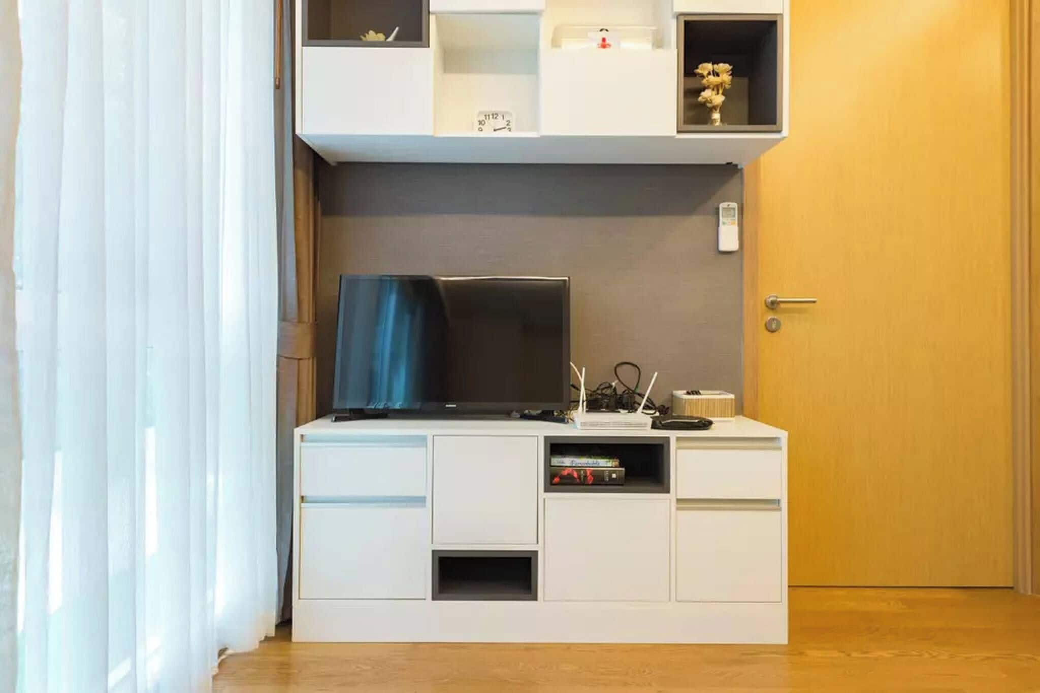 Una habitación con una televisión sobre un mueble blanco, una puerta y una ventana con cortinas.