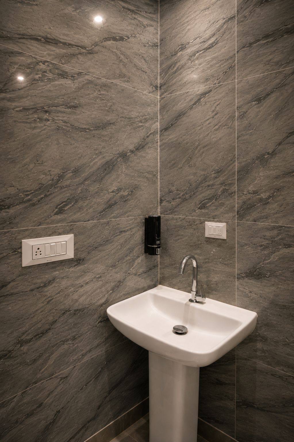 A bathroom with a marble wall, a pedestal sink, a wall-mounted faucet, and a light switch.