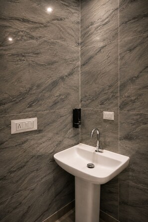 A bathroom with a marble wall, a pedestal sink, a wall-mounted faucet, and a light switch. - Xenious Elite Inn (Greater Noida)