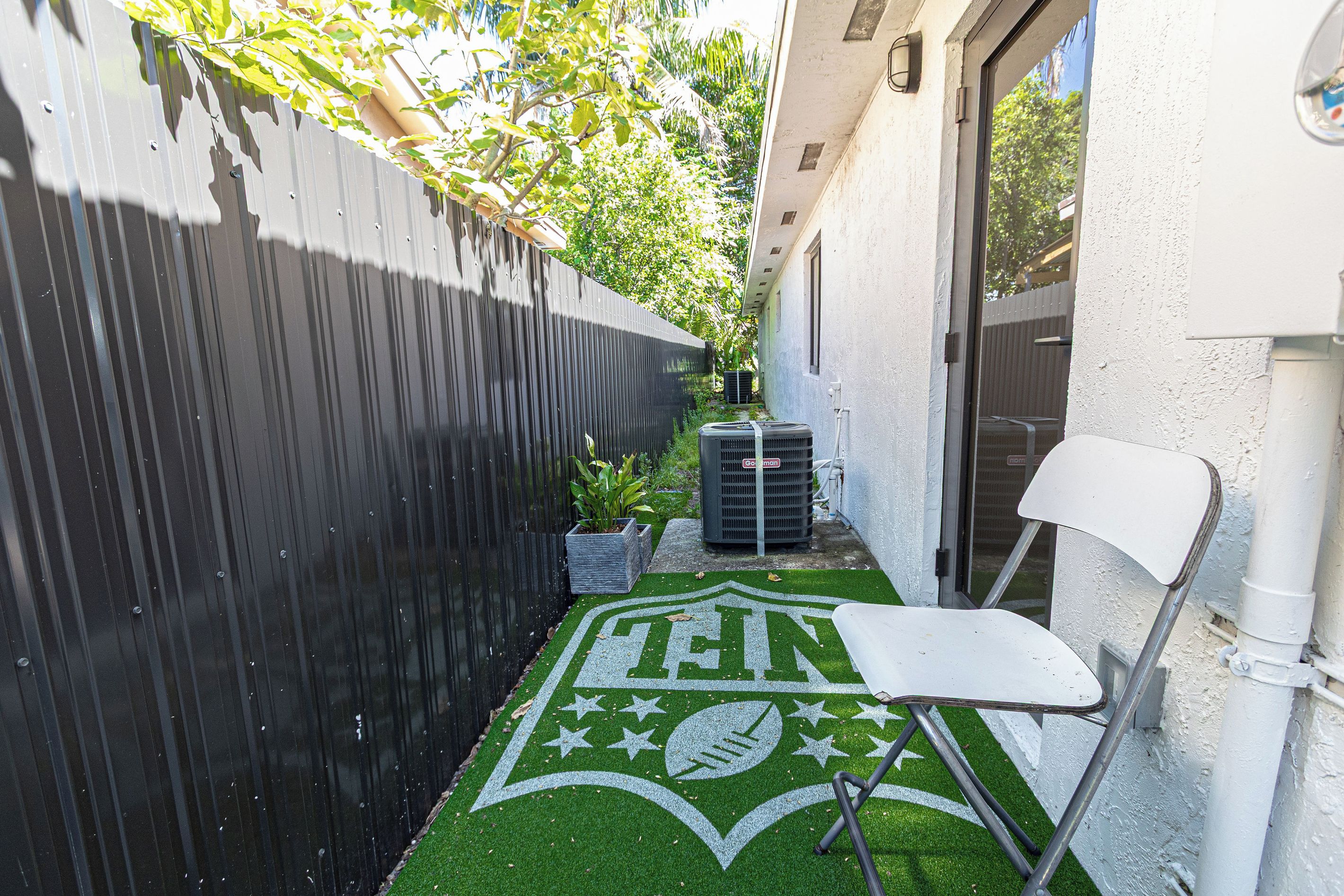 A narrow outdoor space with a green artificial turf area featuring a sports emblem, a white chair, and a black air conditioning unit.