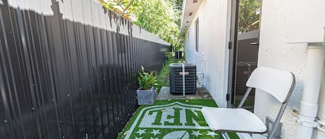 A narrow outdoor space with a green artificial turf area featuring a sports emblem, a white chair, and a black air conditioning unit.