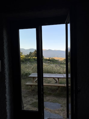 A view through a door with a wooden bench and picnic table outside, overlooking a grassy area and distant mountains. - Maison Aline - Maison familiale avec vue sur les montagnes (Font-Romeu-Odeillo-Via)