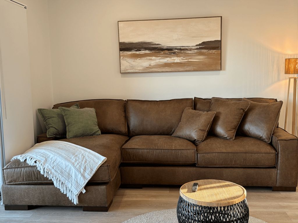 A brown leather sofa with a white throw blanket and a woven stool in a living room with a painting on the wall.