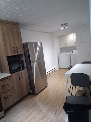A modern kitchen with wooden cabinets, a stainless steel refrigerator, a microwave, a washing machine, and a table with chairs.