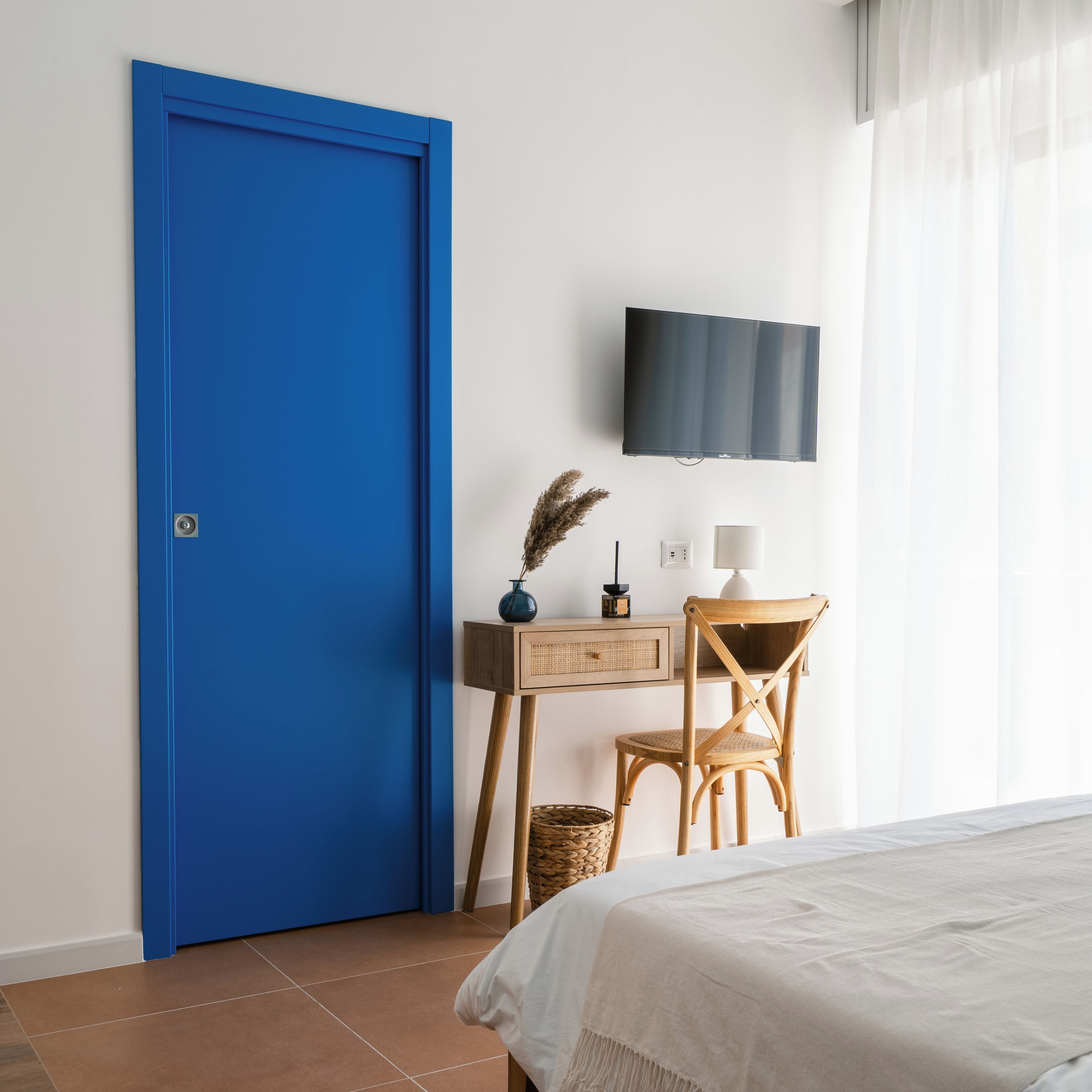 Una stanza con una porta blu, un letto, una scrivania con una sedia, un televisore montato a parete e una finestra con tende.