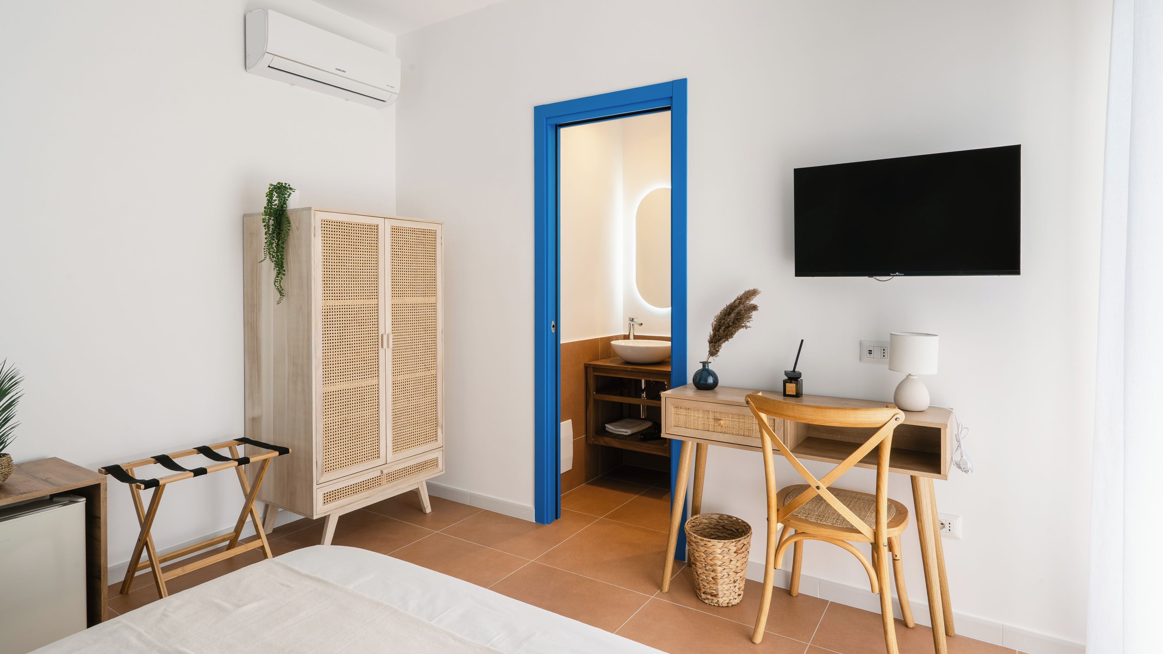 Una habitación de hotel moderna con una cama, un escritorio con una silla, un televisor y un baño visible a través de una puerta azul.