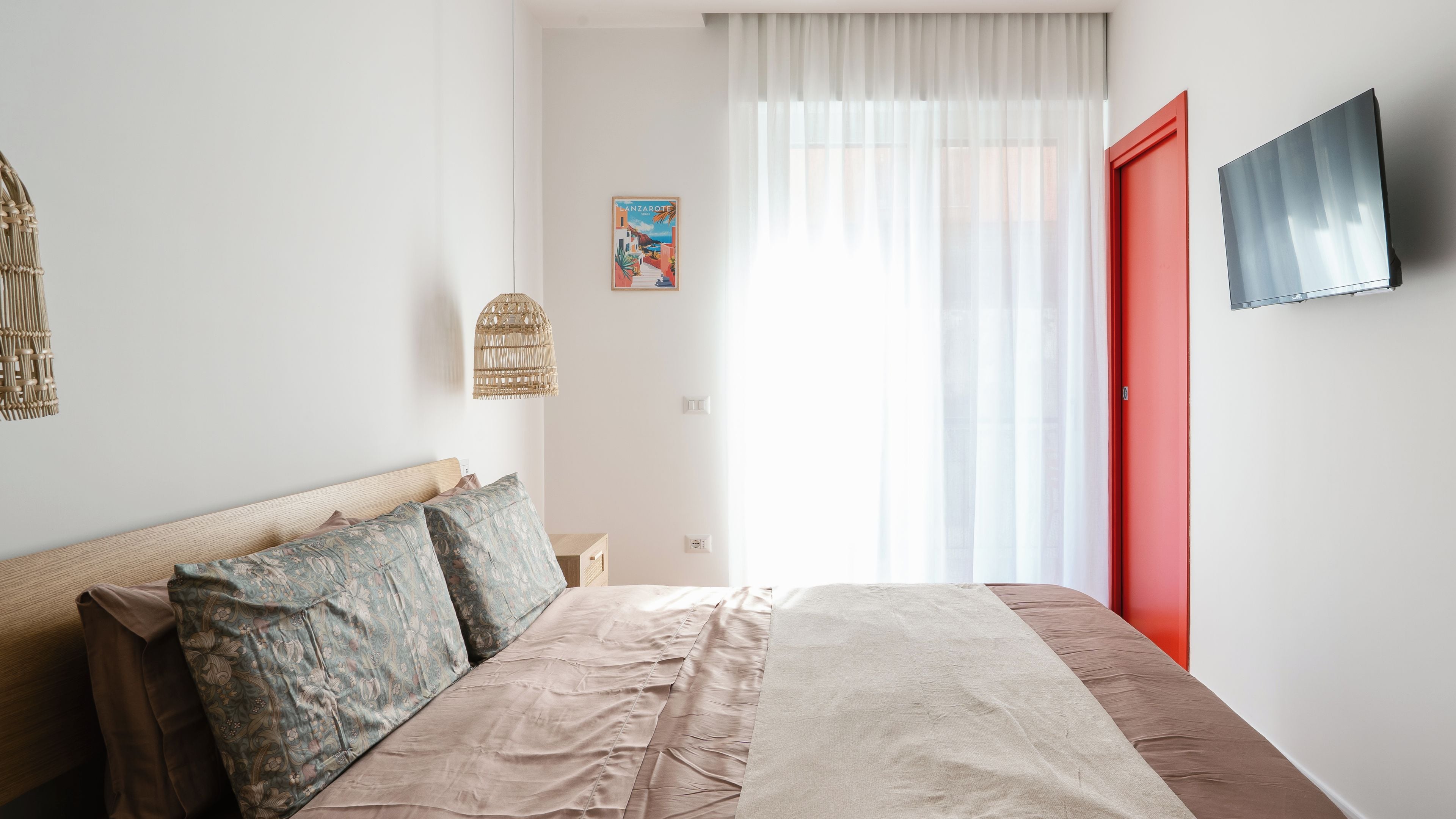 A bedroom with a bed, a wall-mounted TV, a red door, and a window with sheer curtains.