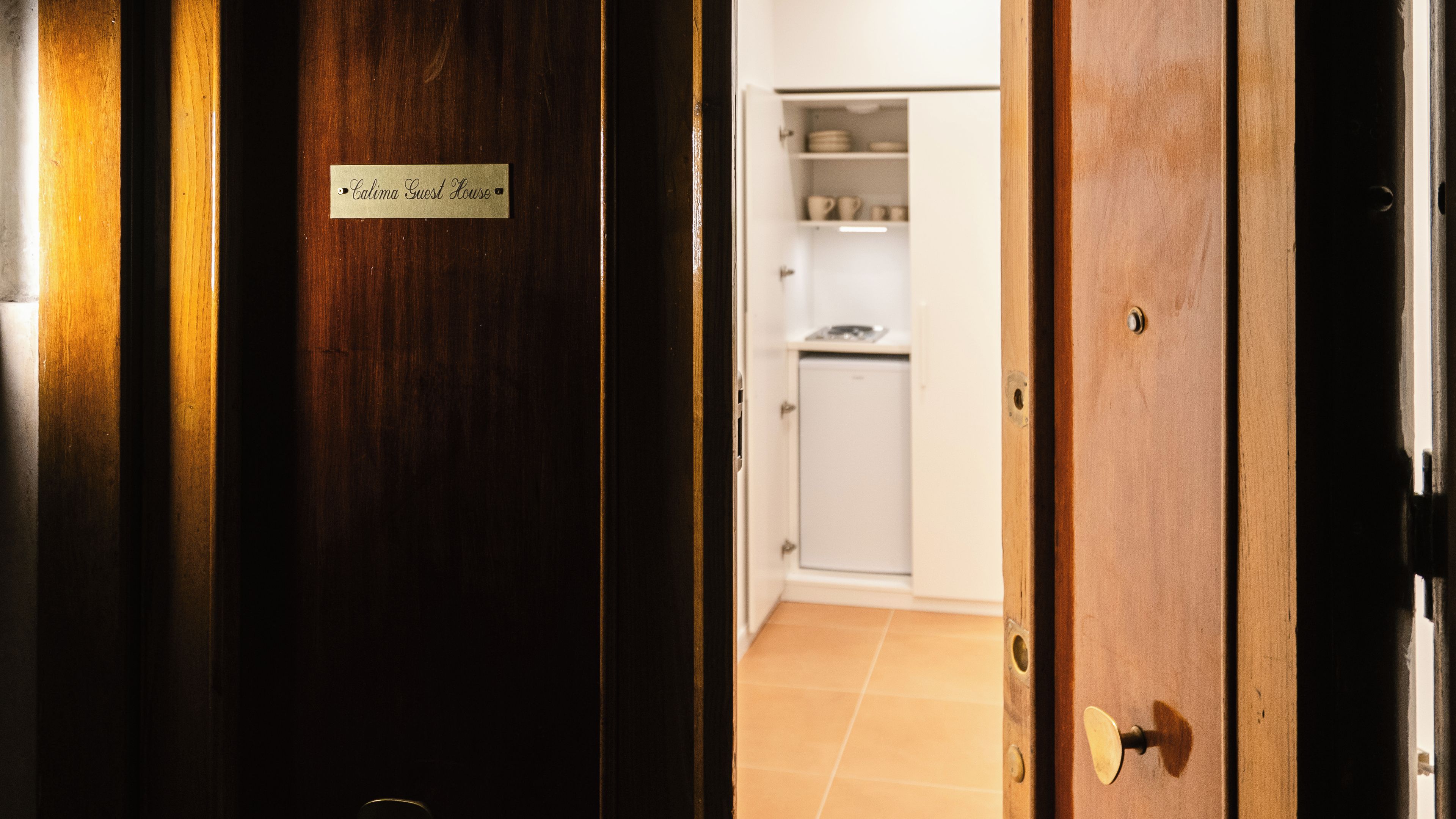 A hotel room with a wooden door, a sign reading 'Calima Guest House,' and a glimpse of a kitchen area with cabinets and a refrigerator.