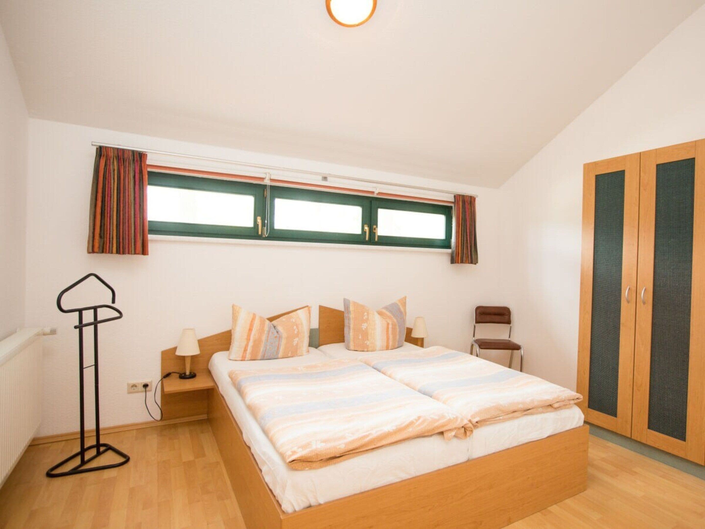 A bedroom with a bed, a chair, a nightstand, a lamp, a window with curtains, and a wardrobe.