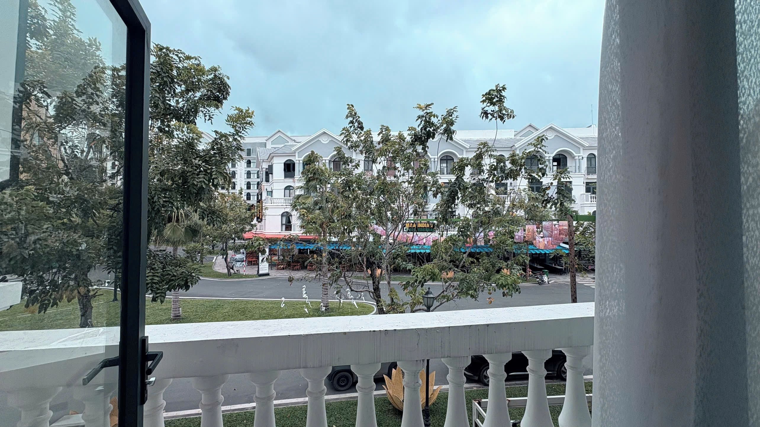 A balcony with a view of a white building with multiple windows and balconies, a street with parked cars, and some greenery.