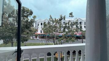 A balcony with a view of a white building with multiple windows and balconies, a street with parked cars, and some greenery.