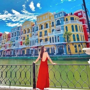 A woman in a red dress standing on a balcony with a view of colorful buildings and a river. - Teddy Pumpkin Homestay-Grand World (Phu Quoc)