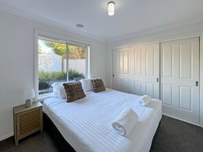A bedroom with a large bed, a window, a bedside table, and a wardrobe. - St Paul's - Sleeps 8 (Wagga Wagga)