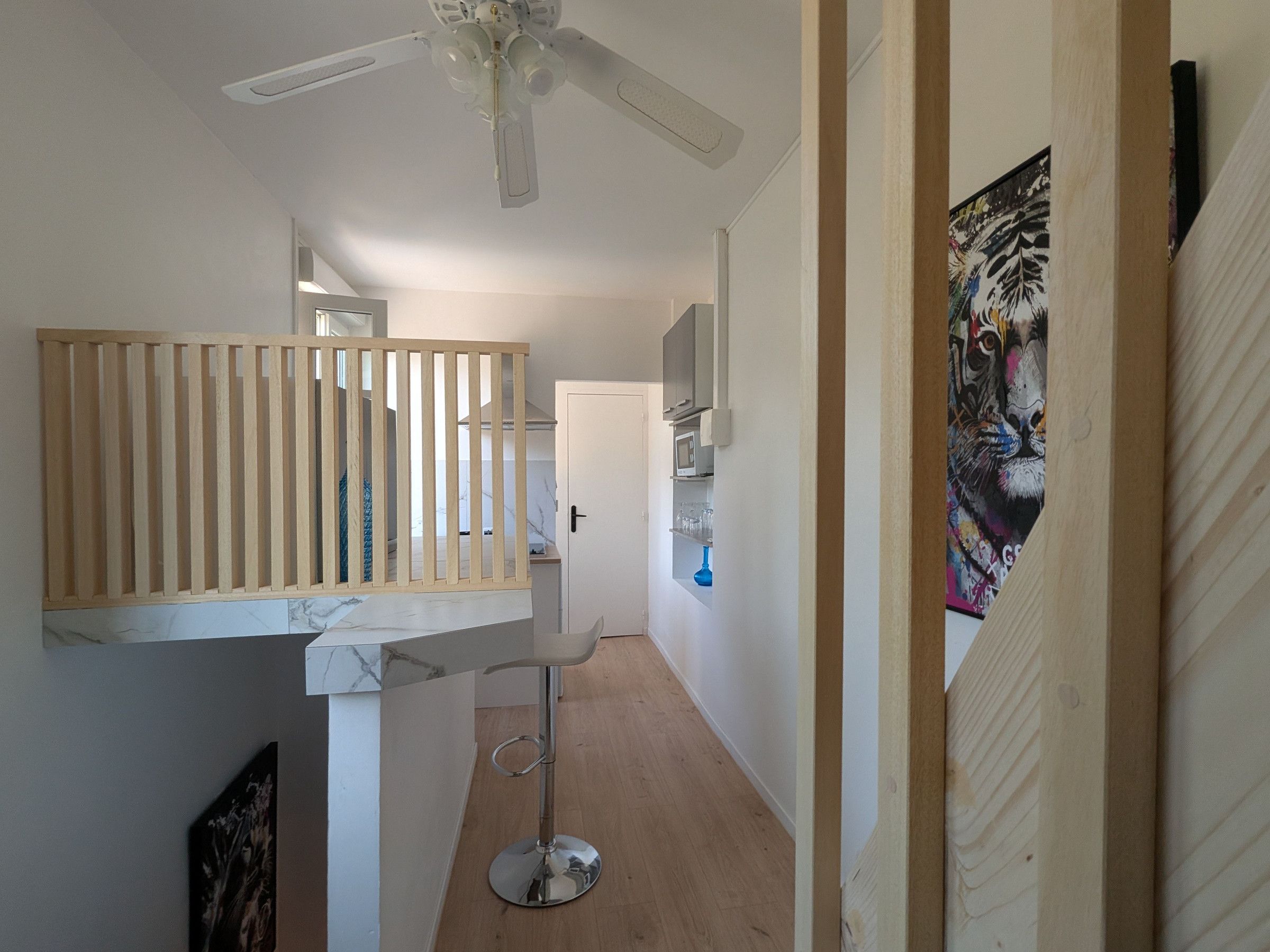 A hallway with a wooden slatted partition, a ceiling fan, a door leading to a kitchen, and a colorful artwork on the wall.
