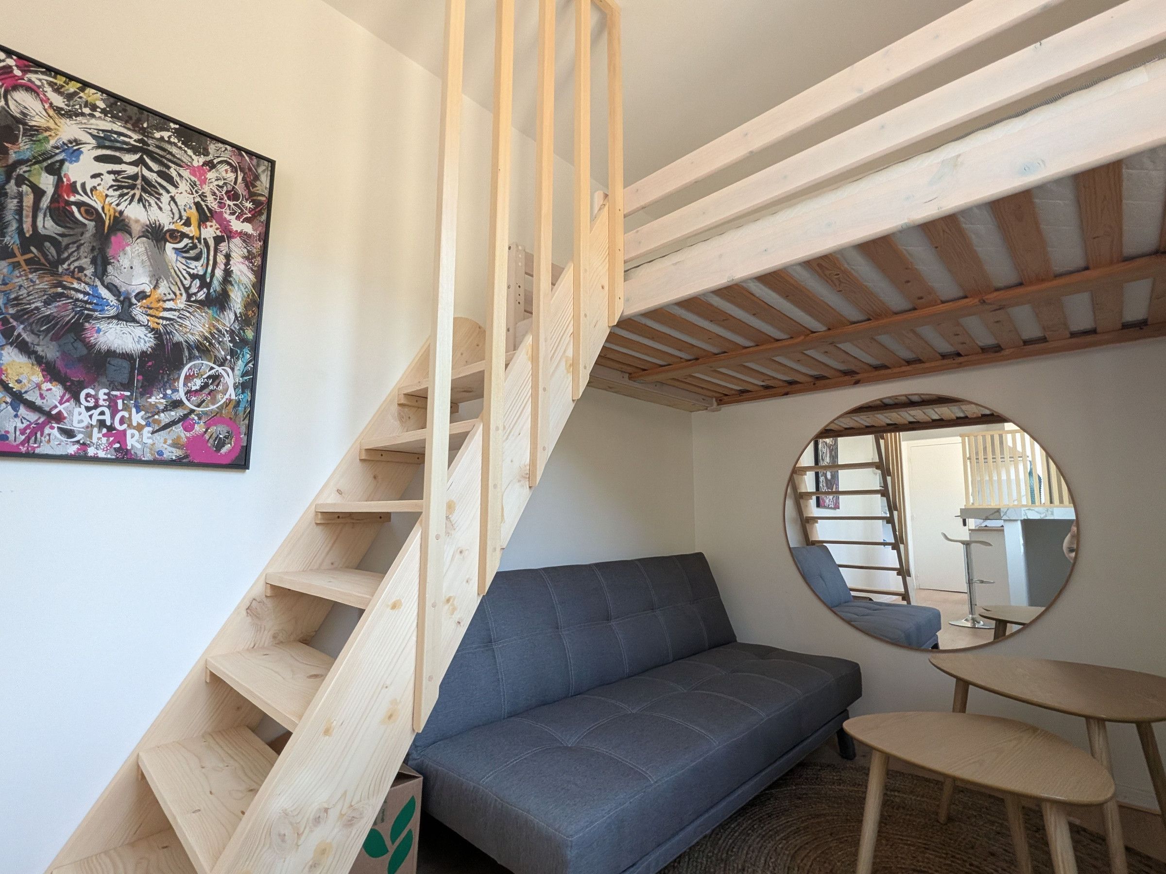 A room with a wooden staircase, a grey sofa, a round mirror, and a painting on the wall.