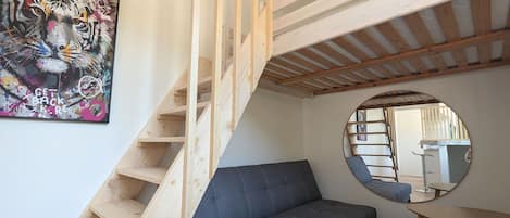 Um quarto com escada de madeira, sofá cinzento, espelho redondo e um quadro na parede.