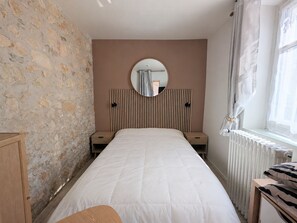 A bedroom with a stone wall, a bed with a white duvet, a round mirror, a radiator, and a window with curtains. - Charming studio in Val de Creuse (Argenton-sur-Creuse)