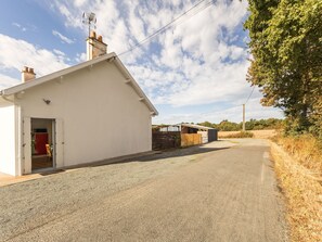 Una carretera rural que lleva a un edificio blanco con puerta roja, una chimenea y un pequeño cobertizo a lo lejos.