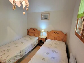 A small room with two wooden beds, a nightstand with a lamp, a framed picture on the wall, and a window with curtains.