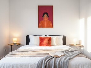 Un letto rifatto con cura, un libro, un cuscino rosso e un quadro incorniciato sopra.