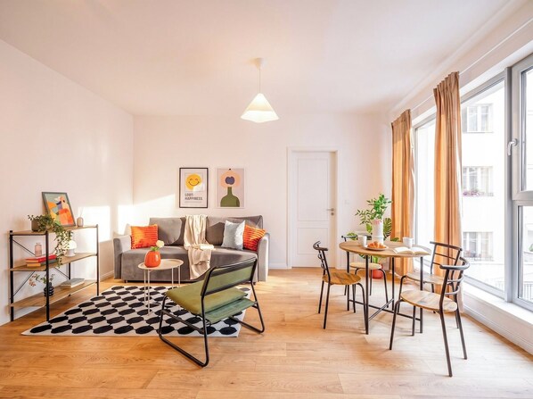 Ein modernes Wohnzimmer mit einer Couch, einem Couchtisch und Sesseln, ein Essbereich mit Tisch und Stühlen sowie große Fenster mit Vorhängen.