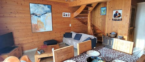 L’intérieur chaleureux d’un chalet, avec un revêtement mural en bois, une table à manger dressée avec des assiettes et des verres, un canapé et un escalier.