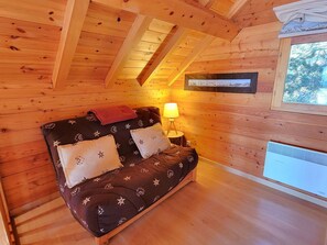 Acogedor cuarto de cabaña de madera con cama, mesita de noche, lámpara y ventana con vistas a árboles.
