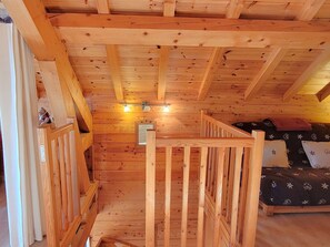 Interior de una cabaña de madera con una cama en el entrepiso, una pequeña escalera y barandilla de madera.