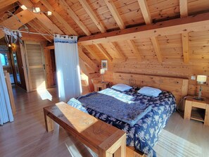 Un dormitorio acogedor en el ático, con techo de madera, una cama con ropa de cama estampada, un banco y una mesita de noche con lámpara.