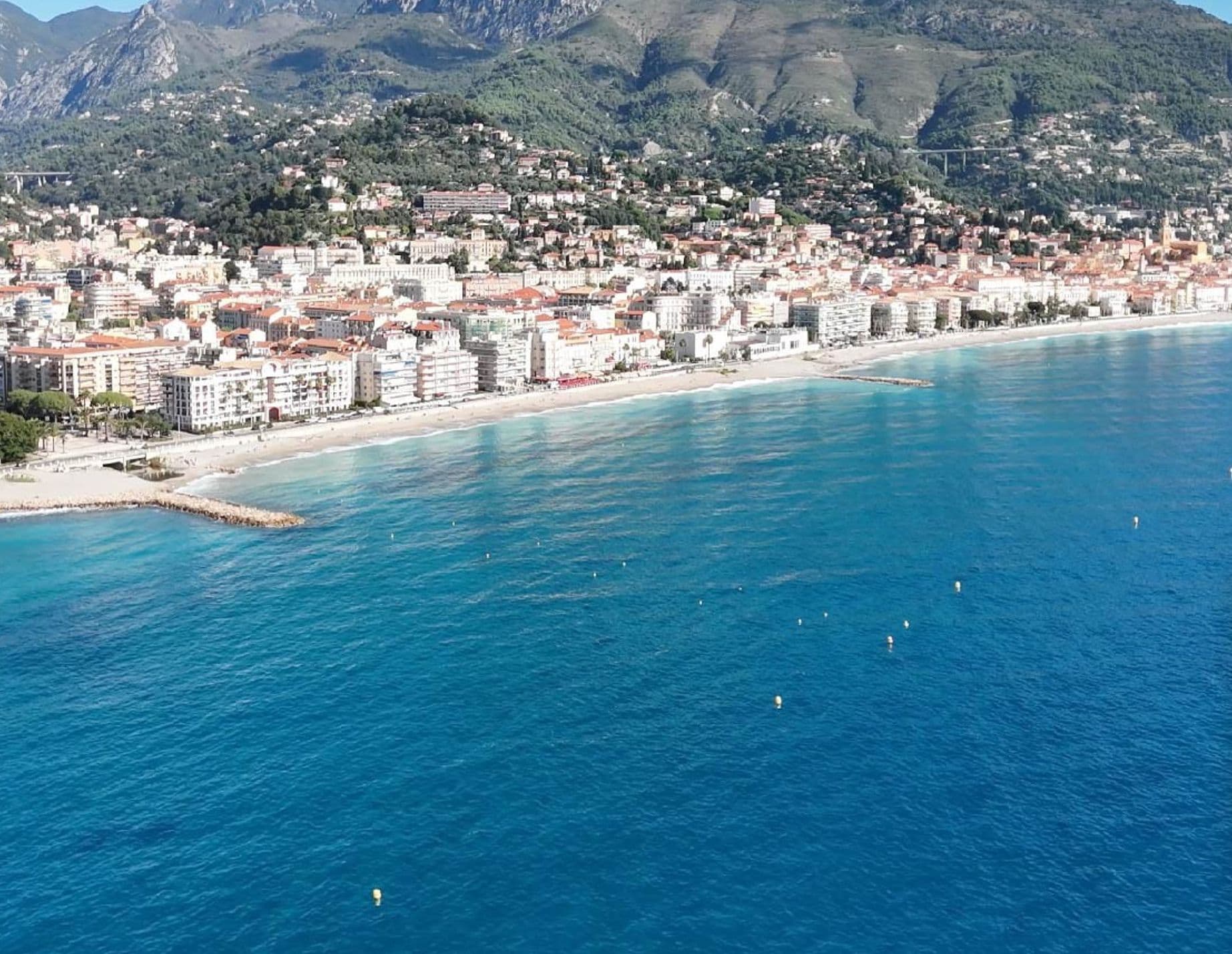 A coastal cityscape with buildings along the waterfront, a clear blue sea, and mountains in the background.