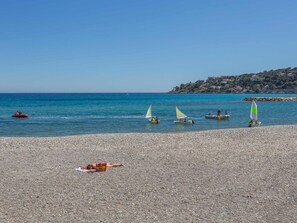 A pebbled beach with several sailboats on the water, a distant shoreline with buildings, and a person sunbathing. - Apartment with AC and parking facing the sea (Roquebrune-Cap-Martin)